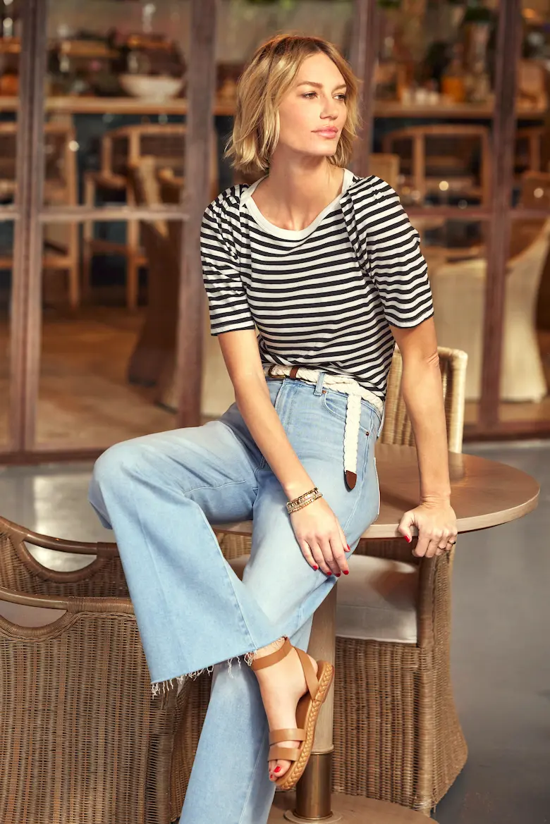 A woman with short blonde hair wearing an outfit consisting of a striped top and wide-leg jeans sits casually on a wooden table. This wide leg jeans outfit exemplifies casual yet stylish dressing.